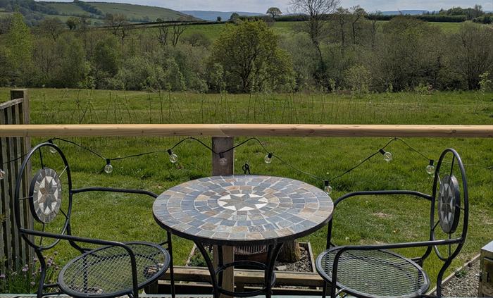 Metal and stone mosaic outdoor table and chairs on the wood deck with a view over the wildflower meadow to the trees round the lake and the hills beyo